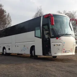 Biały autokar z otwartymi drzwiami na zewnątrz, ozdobiony kolorowymi gwiazdkami, zaparkowany na żwirowym terenie z drzewami w tle.