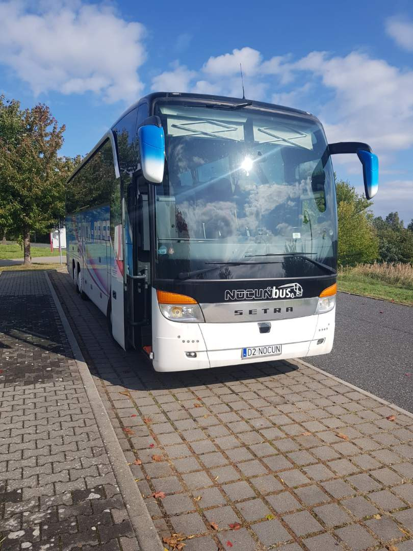 Biały autokar turystyczny Setra z niebieskimi lusterkami, zaparkowany na brukowanym parkingu w słoneczny dzień, widoczne logo Noctunbus.