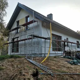 Dom jednorodzinny w trakcie prac elewacyjnych, z widocznym rusztowaniem, częściowo ocieploną ścianą i żółtym wężem wentylacyjnym, na pierwszym planie ziemia i elementy konstrukcyjne.