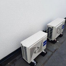 Lomachłód Leszek Steinert - Dwa białe, zewnętrzne moduły klimatyzacji Haier ustawione na czarnych podstawkach na zewnątrz budynku. Widoczna biała, teksturowana ściana budynku w tle.