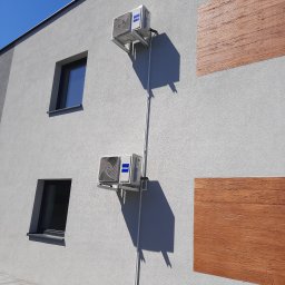 Lomachłód Leszek Steinert - Dwa klimatyzatory Haier zamontowane na szarej ścianie budynku z oknami i drewnopodobnymi panelami dekoracyjnymi, widoczne rury odprowadzające skropliny.