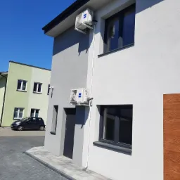 Dwa klimatyzatory marki Haier zamontowane na elewacji dwukondygnacyjnego budynku mieszkalnego w słoneczny dzień, widok z poziomu chodnika.