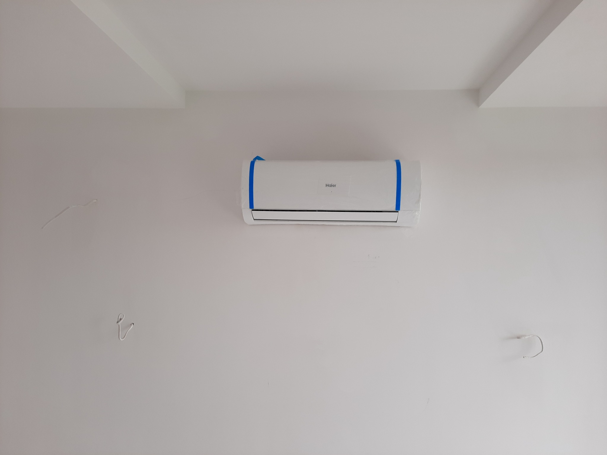 Nowo zamontowana, biała klimatyzacja Haier na białej ścianie, zabezpieczona niebieską folią ochronną, widoczne ślady po przewodach.