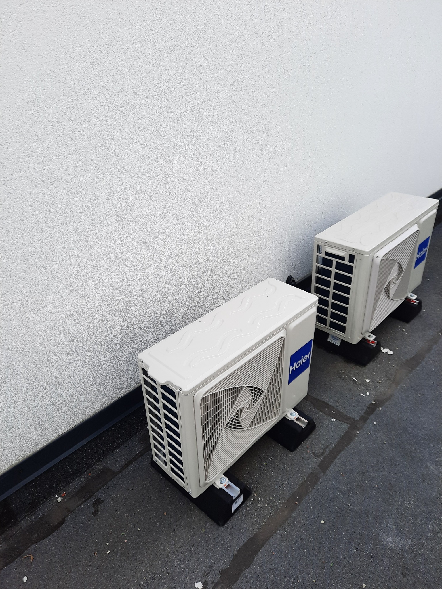 Dwa białe, zewnętrzne moduły klimatyzacji Haier ustawione na czarnych podstawkach na zewnątrz budynku. Widoczna biała, teksturowana ściana budynku w tle.
