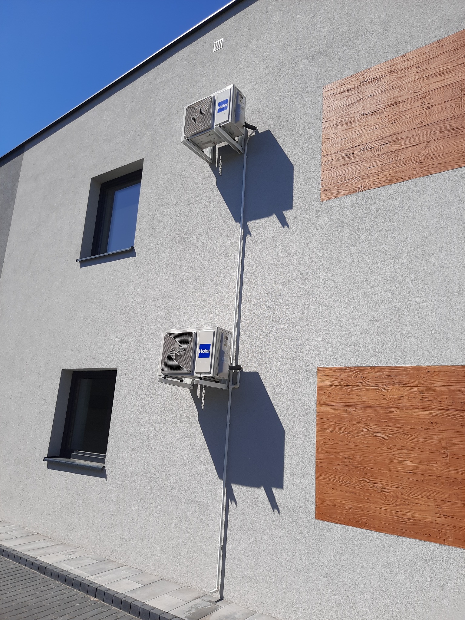 Dwa klimatyzatory Haier zamontowane na szarej ścianie budynku z oknami i drewnopodobnymi panelami dekoracyjnymi, widoczne rury odprowadzające skropliny.