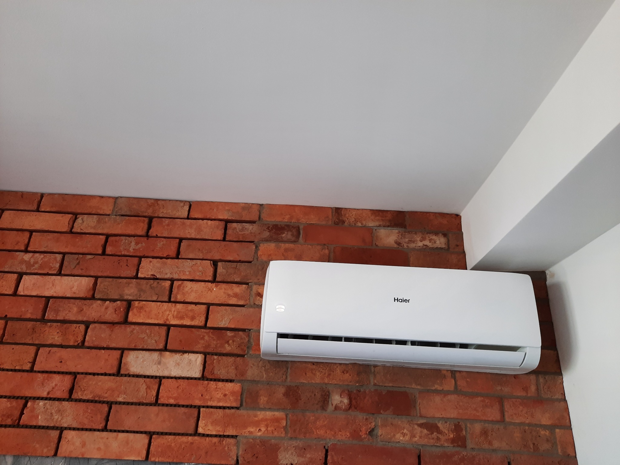 Biały klimatyzator Haier zamontowany na ceglanej ścianie w pomieszczeniu z białym sufitem.