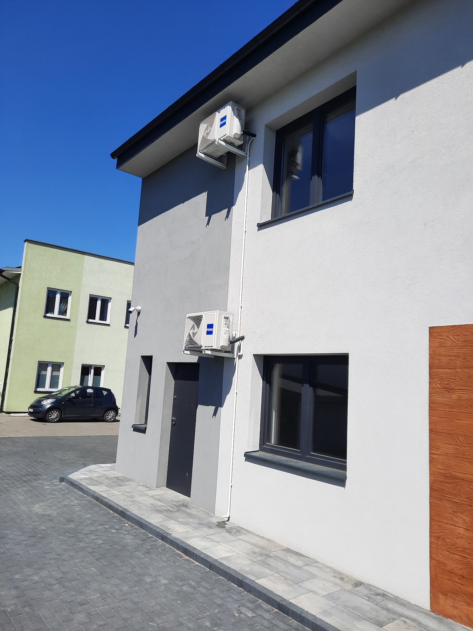 Dwa klimatyzatory marki Haier zamontowane na elewacji dwukondygnacyjnego budynku mieszkalnego w słoneczny dzień, widok z poziomu chodnika.