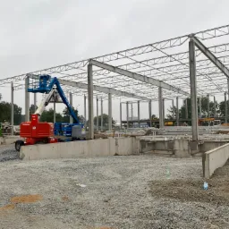 Wznoszenie szkieletu hali: widok na konstrukcję stalową z betonowymi fundamentami, podnośniki koszowe Genie i Mateco na placu budowy, pochmurne niebo w tle.