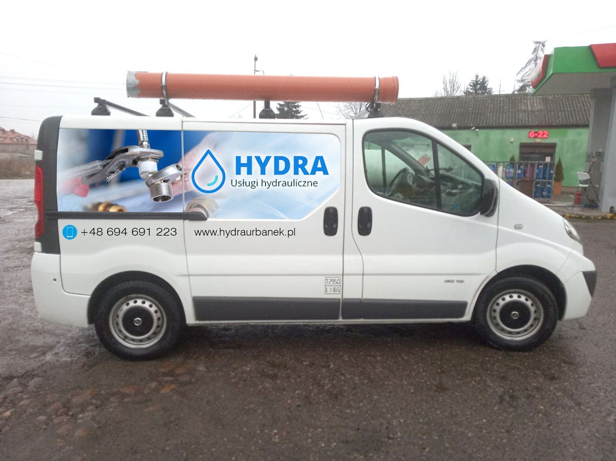 Biały samochód dostawczy z reklamą firmy hydraulicznej Hydra, zaparkowany na mokrym asfalcie przed budynkiem z zielonym dachem i czerwonym napisem 6-22. Na dachu samochodu widoczna rura PCV.