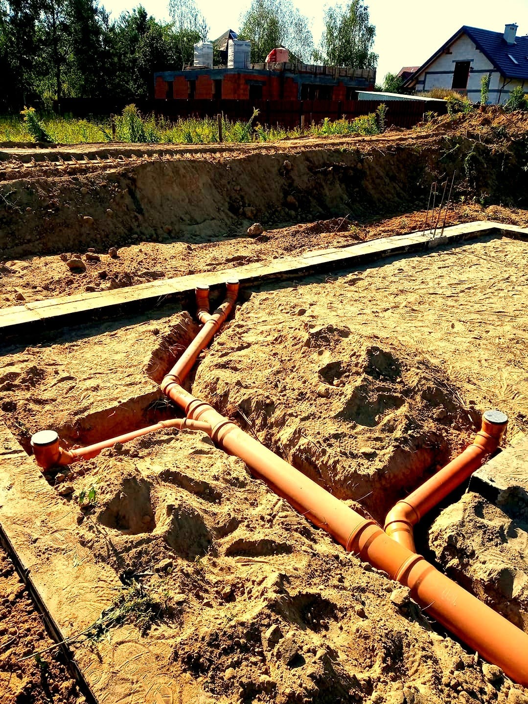 Pomarańczowe rury kanalizacyjne ułożone w wykopie z piasku, widoczne kształtki i zakończenia z dekielkami, w tle fundamenty budynków w trakcie budowy.