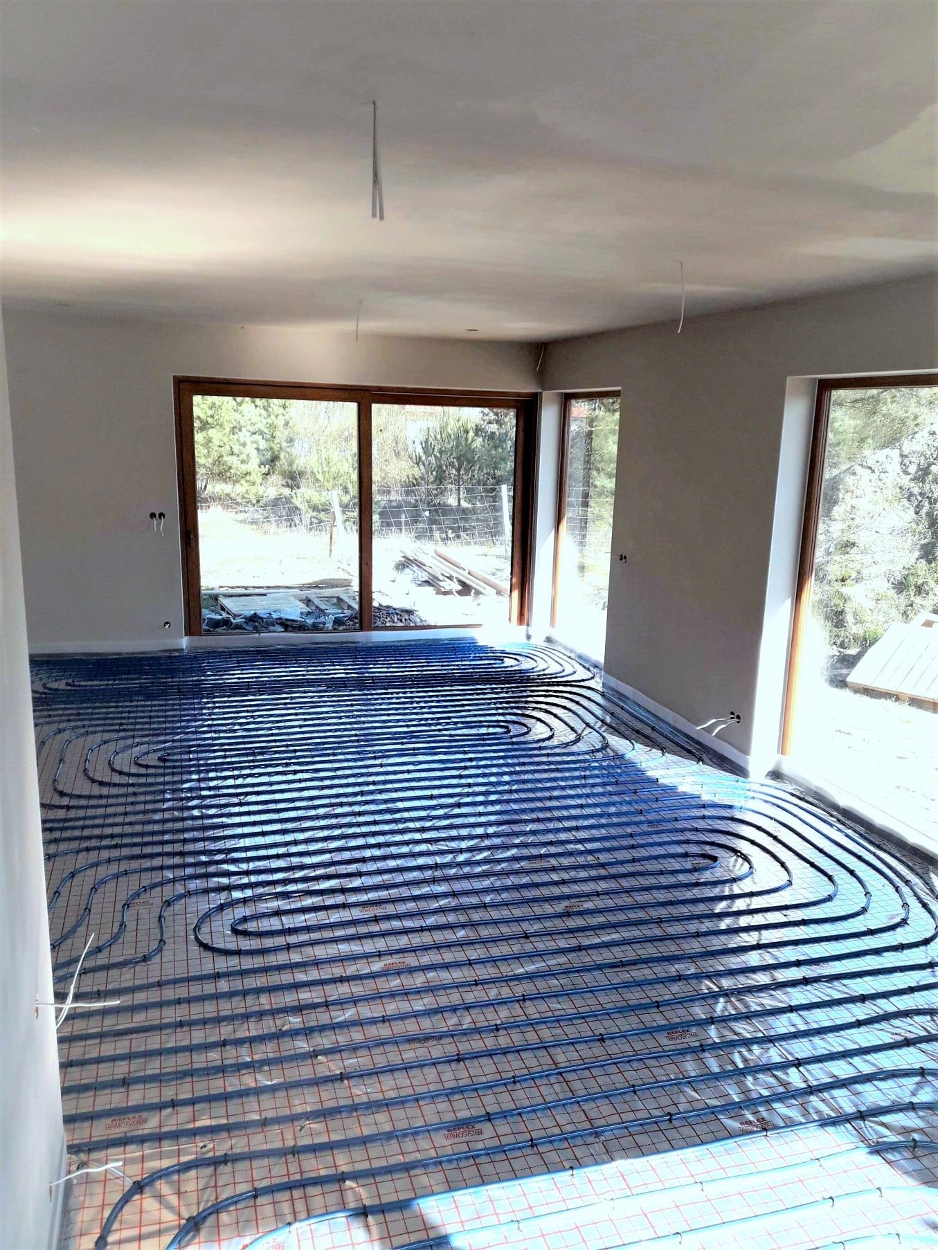 Niebieskie rurki ogrzewania podłogowego ułożone na izolacji z siatką w jasnym, przestronnym pokoju z dużymi oknami wychodzącymi na zieleń.