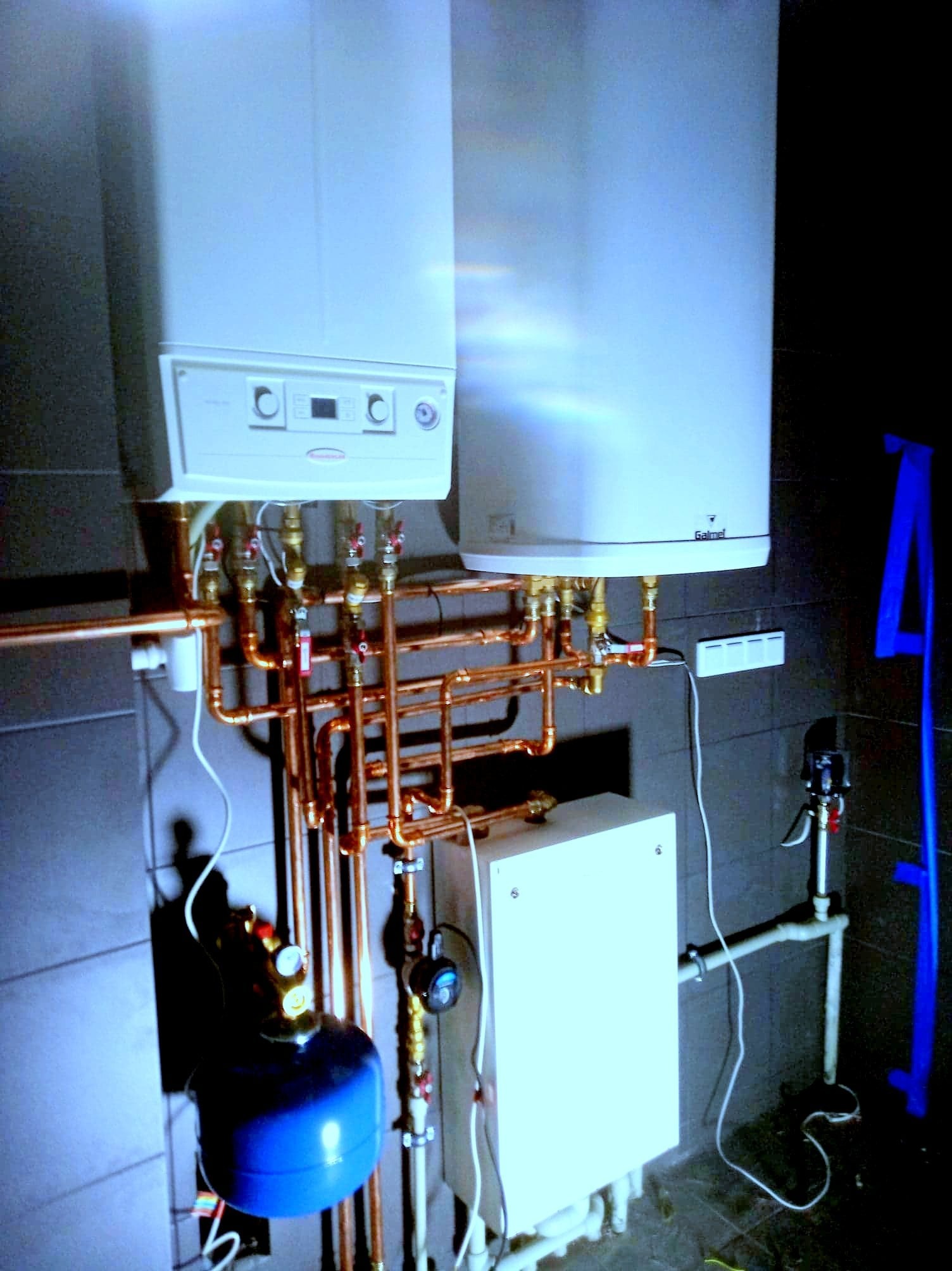 Nowoczesny dwufunkcyjny piec gazowy z miedzianą instalacją, widoczny niebieski zbiornik wyrównawczy i biała skrzynka sterownicza na tle ciemnych, płytkowych ścian.