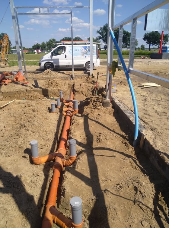 Pomarańczowe rury kanalizacyjne z szarymi zakończeniami ułożone w wykopie ziemnym na placu budowy, widoczny fragment metalowej konstrukcji i samochód dostawczy z logiem firmy w tle.