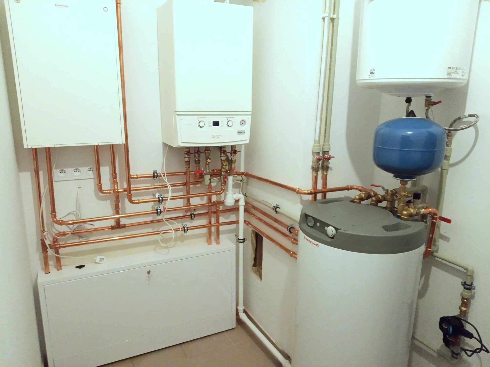 Zestaw urządzeń grzewczych: dwa piece gazowe, zasobnik ciepłej wody z niebieskim naczyniem przeponowym, połączone miedzianymi rurami w kotłowni. Widoczne liczne zawory i przyłącza.