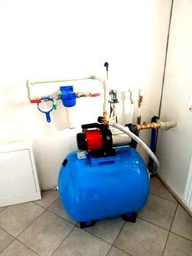 Niebieski hydrofor z osprzętem (filtry, rury) zamontowany w pomieszczeniu z białymi ścianami i szarą podłogą z płytek. Widoczny fragment grzejnika.