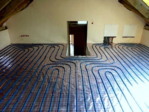 Montaż rur systemu grzewczego podłogowego na poddaszu, widoczne niebieskie rury ułożone w pętle na folii z siatką, surowe ściany i drewniana konstrukcja dachu.