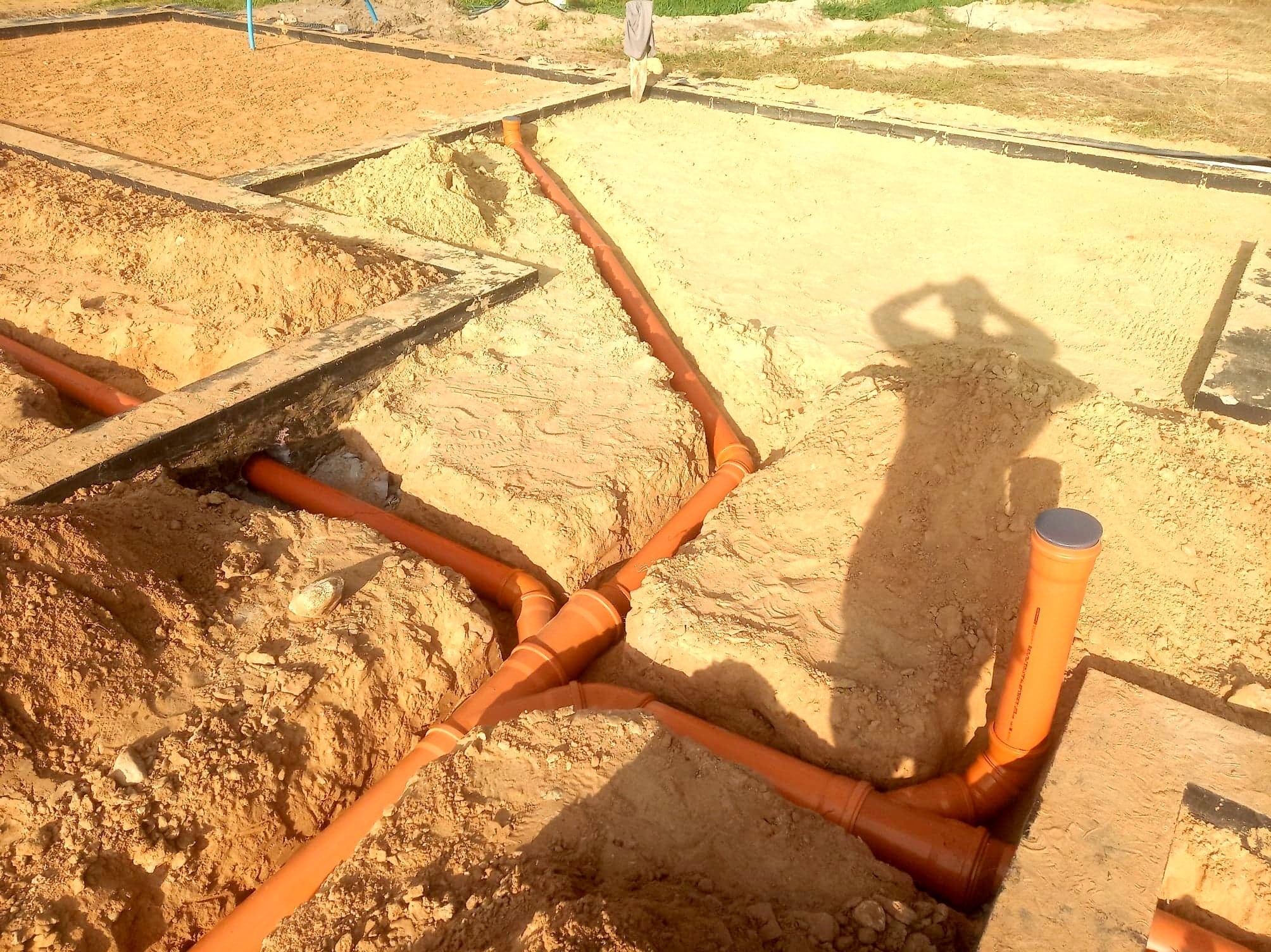 Pomarańczowe rury kanalizacyjne ułożone w wykopie ziemnym na działce budowlanej, widoczny cień osoby robiącej zdjęcie.