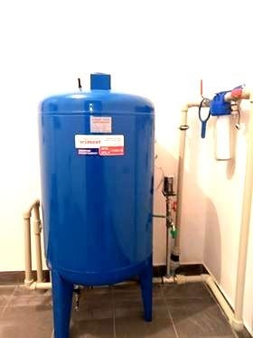 Niebieski zbiornik hydroforowy z widocznymi rurami i manometrem, zamontowany przy białej ścianie w pomieszczeniu z szarą podłogą. Na ścianie widoczny również filtr do wody z niebieskim zaworem.