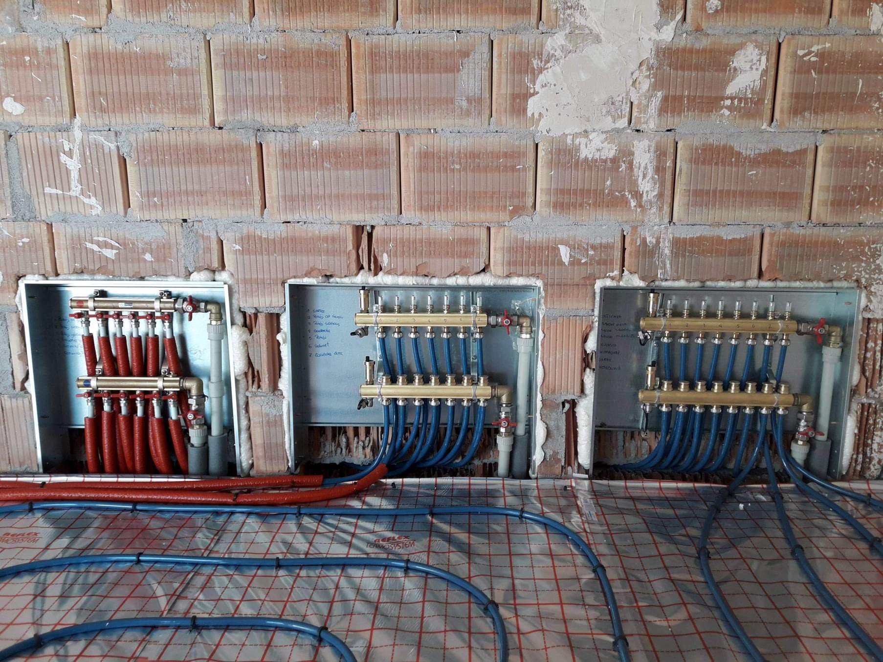 Rozdzielacze ogrzewania podłogowego, jeden z czerwonymi rurami, dwa z niebieskimi, zamontowane w ścianie z cegły dziurawki, instalacja w trakcie montażu na folii z nadrukiem.