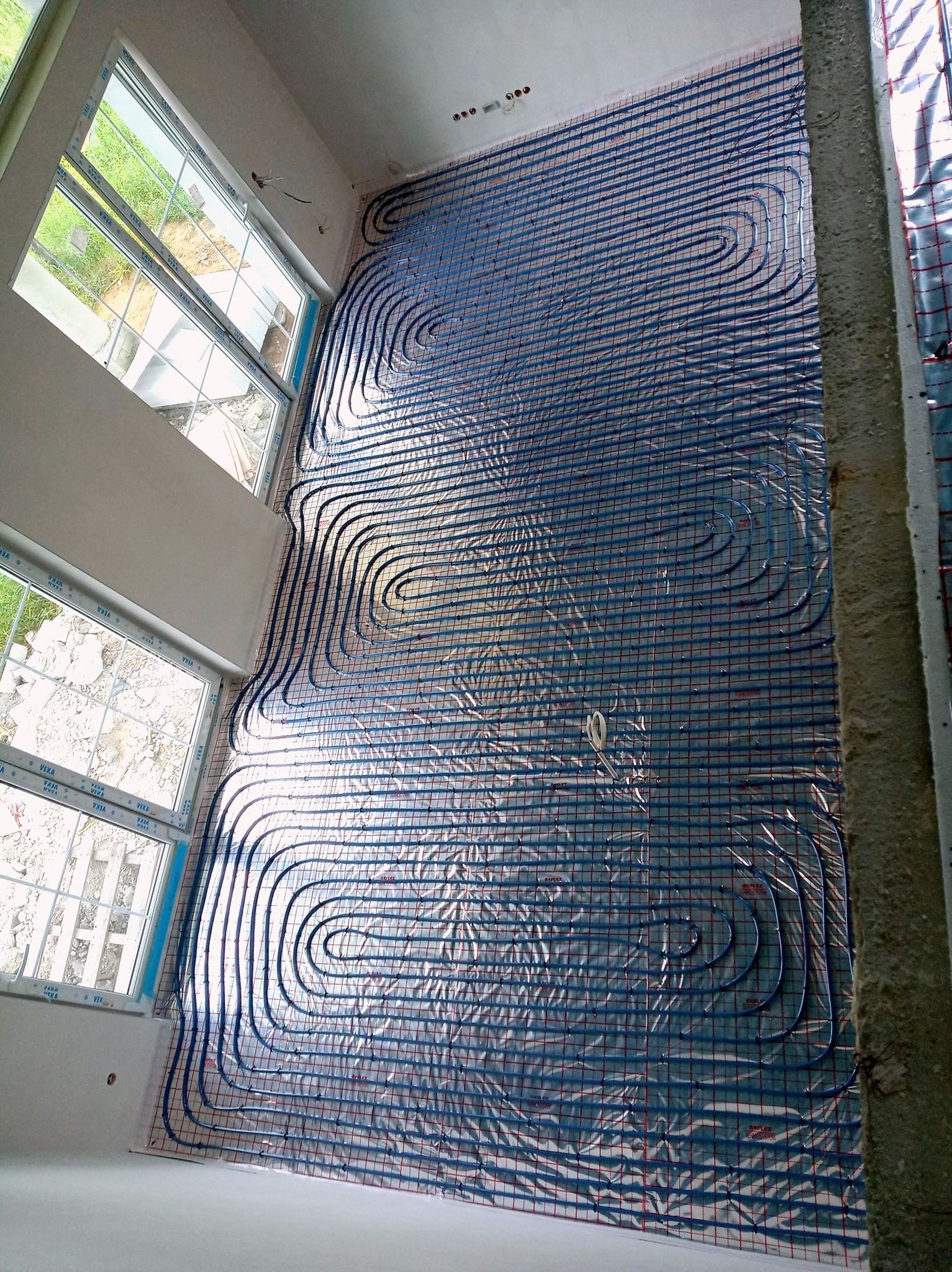 Rozłożona instalacja ogrzewania podłogowego z niebieskich rurek na folii refleksyjnej w surowym wnętrzu z oknami.