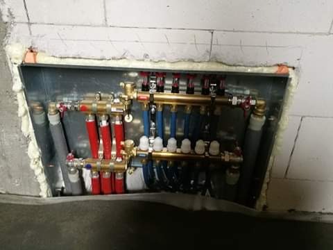 Rozdzielacz ogrzewania podłogowego zamontowany w metalowej skrzynce w ścianie z betonu komórkowego, uszczelniony pianką montażową. Widoczne rury doprowadzające i odprowadzające wodę, zawory...