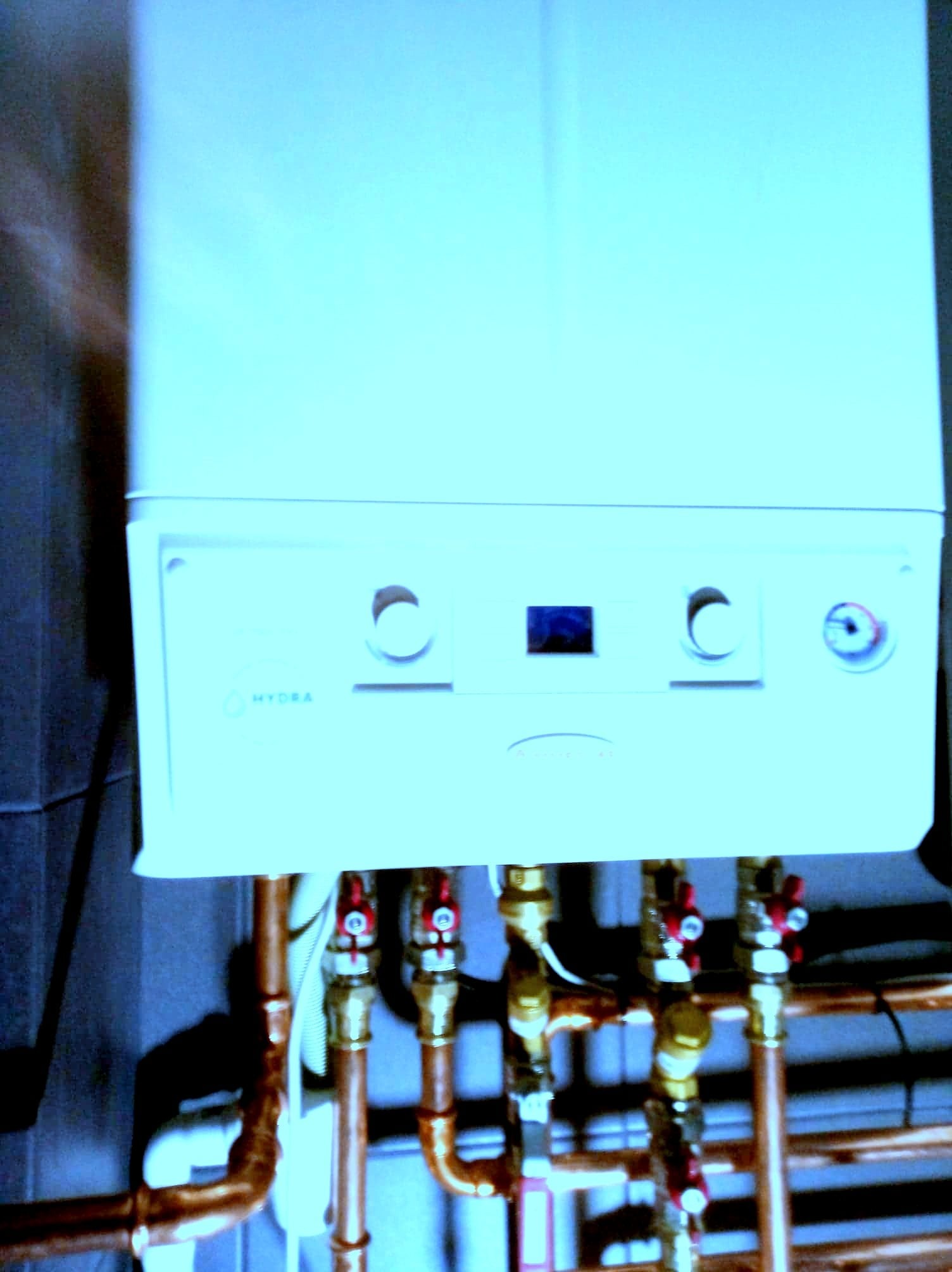 Biały piec gazowy marki Hydra z miedzianymi rurami i czerwonymi zaworami, zamontowany na ciemnej ścianie.