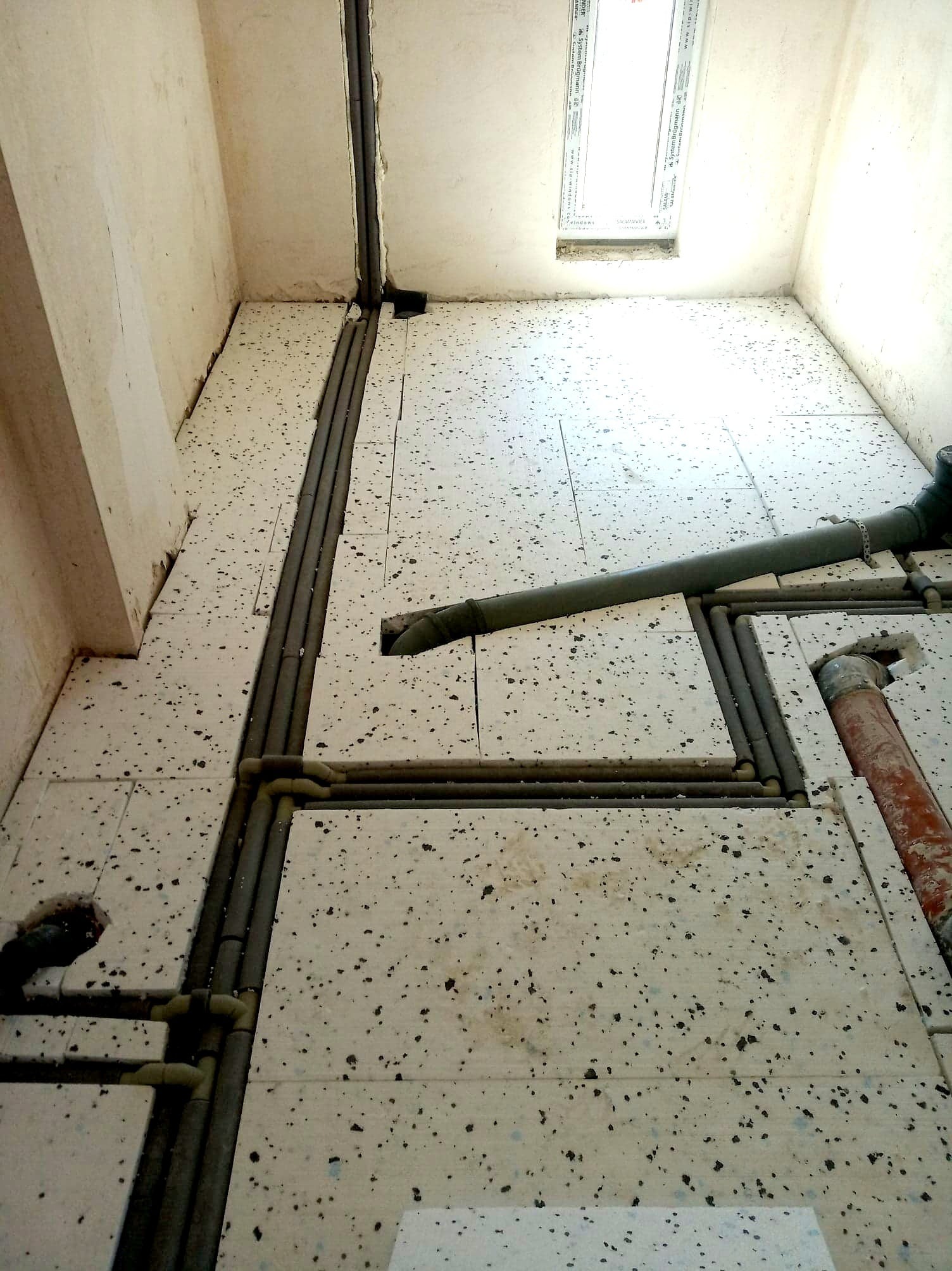 Montaż rur w izolacji podłogowej w surowym wnętrzu, widoczne fragmenty instalacji wodnej i kanalizacyjnej, rury poprowadzone w wycięciach styropianu pod wylewkę.