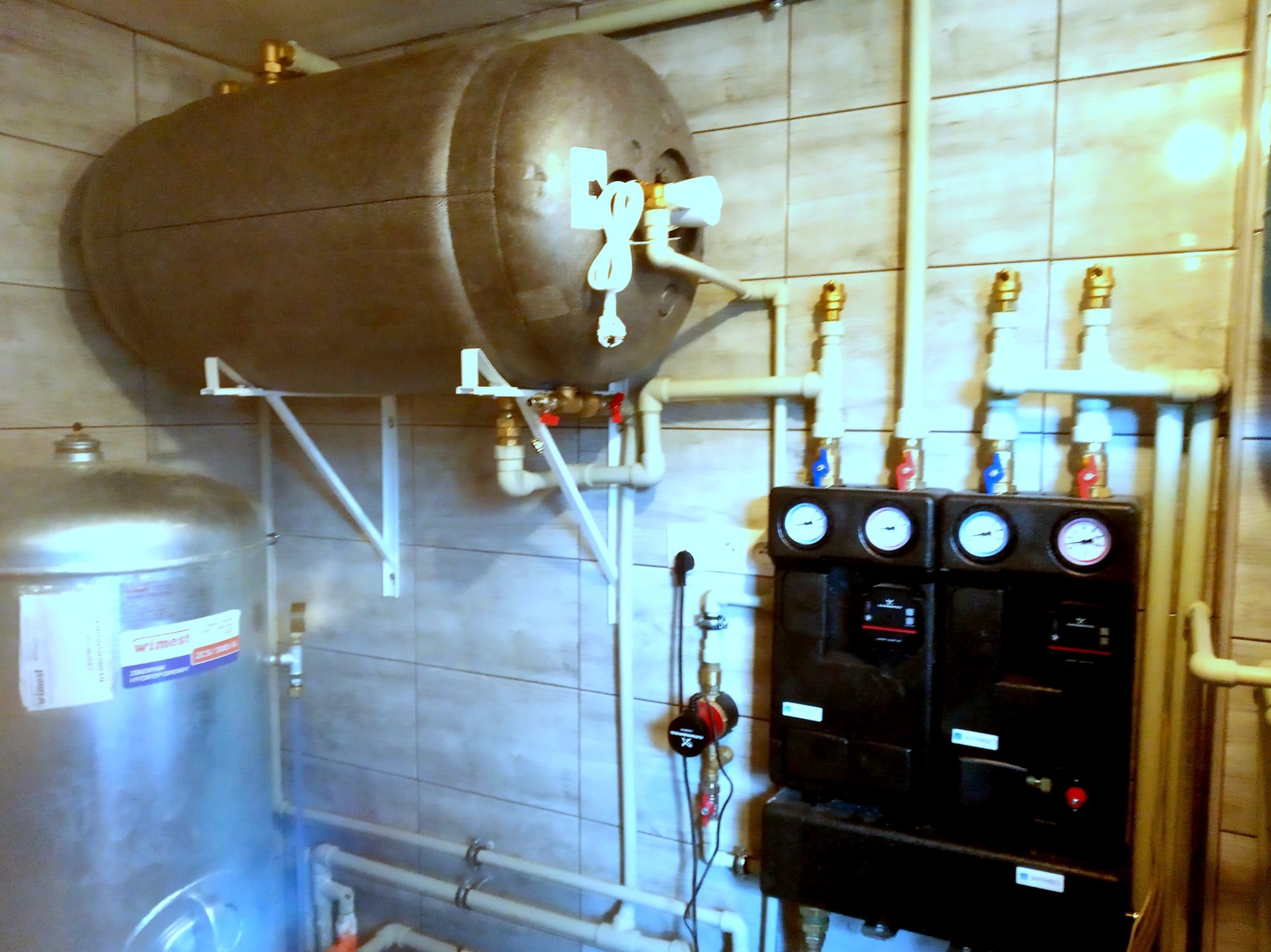 Szary zbiornik akumulacyjny ciepła zawieszony na białych wspornikach, z widocznymi rurami, zaworami i czarnym rozdzielaczem z manometrami na tle płytek ceramicznych.