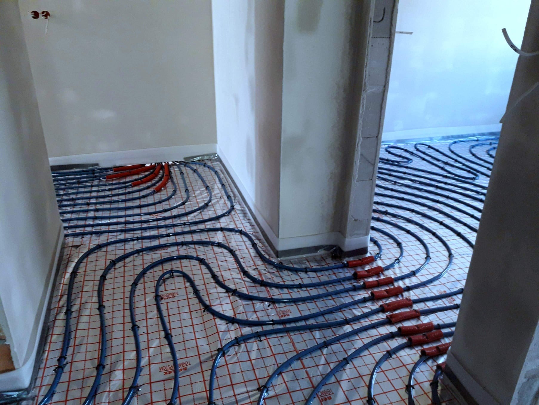 Rozłożone rury ogrzewania podłogowego na siatce, widoczne czerwone złączki i surowe ściany w trakcie remontu.