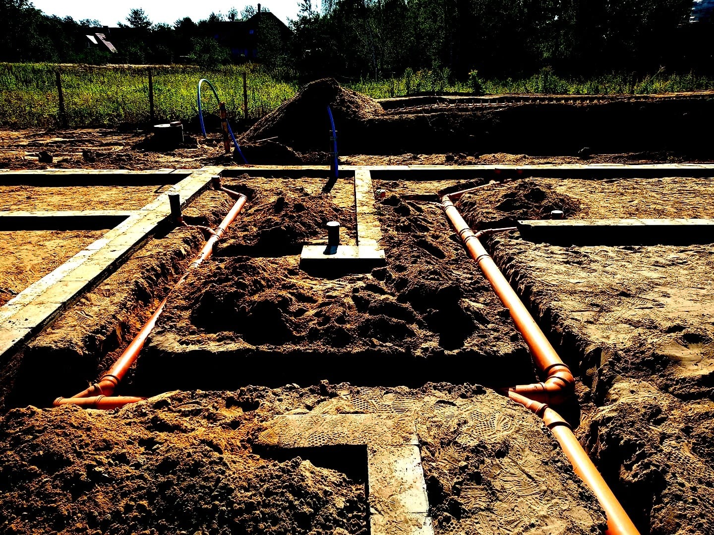 Wykop pod fundamenty z ułożonymi pomarańczowymi rurami kanalizacyjnymi i niebieskimi rurami doprowadzającymi wodę na placu budowy.