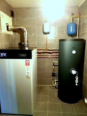 Kocioł grzewczy z czarnym zasobnikiem ciepłej wody w kotłowni wyłożonej szarymi płytkami. Widoczne rury miedziane i dwa zbiorniki wyrównawcze ciśnienia w kolorze niebieskim i białym.