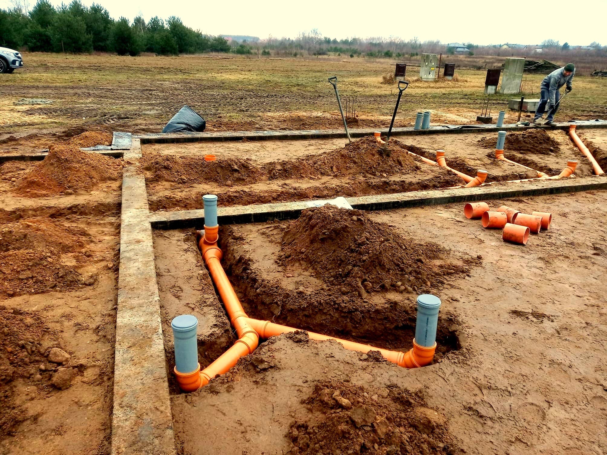 Montaż pomarańczowych rur kanalizacyjnych w wykopie na placu budowy, widoczne szalunki fundamentów, ziemia i pracownik z łopatą w tle.