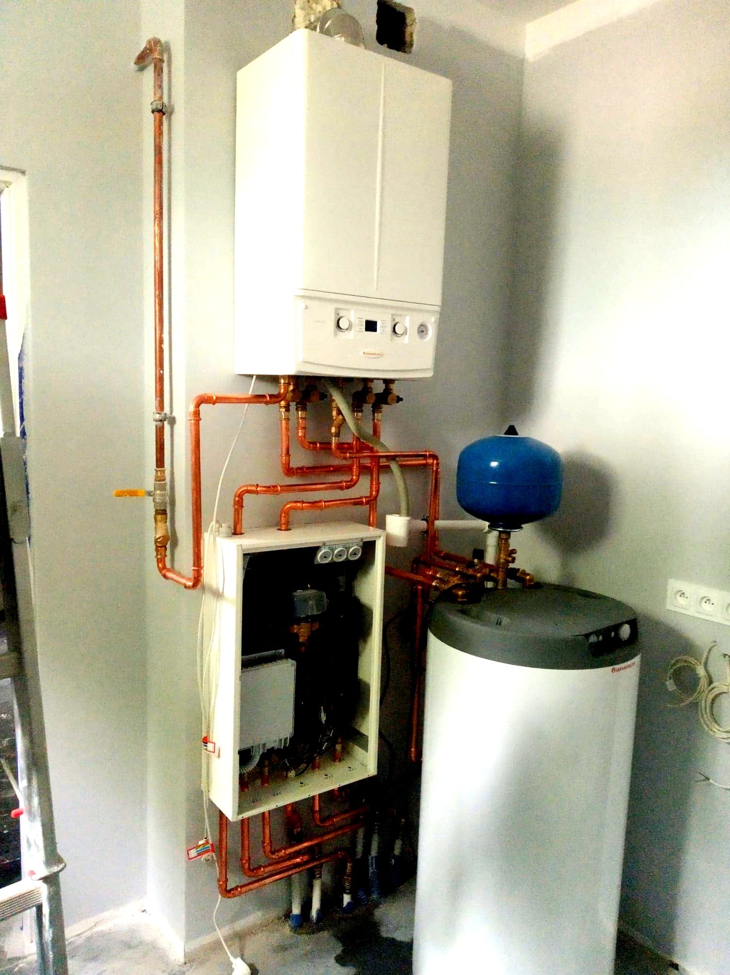 Biały piec gazowy kondensacyjny z miedzianymi rurami i niebieskim naczyniem wzbiorczym, zamontowany na szarej ścianie z widoczną instalacją elektryczną i fragmentem drabiny.