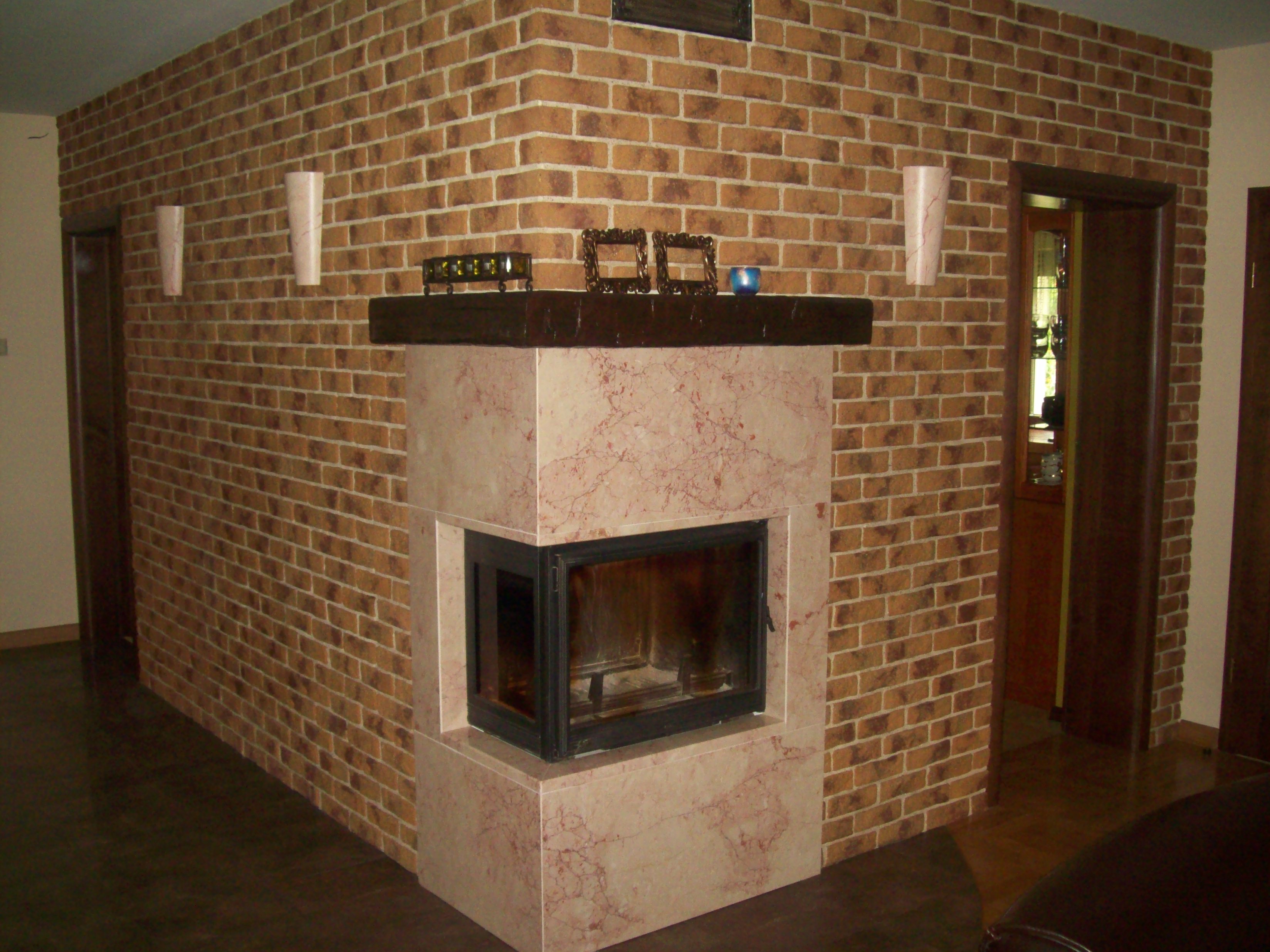 Wnętrze pokoju z kominkiem obudowanym marmurem, ściany wykończone cegłą klinkierową, na kominku dekoracje i oświetlenie ścienne.