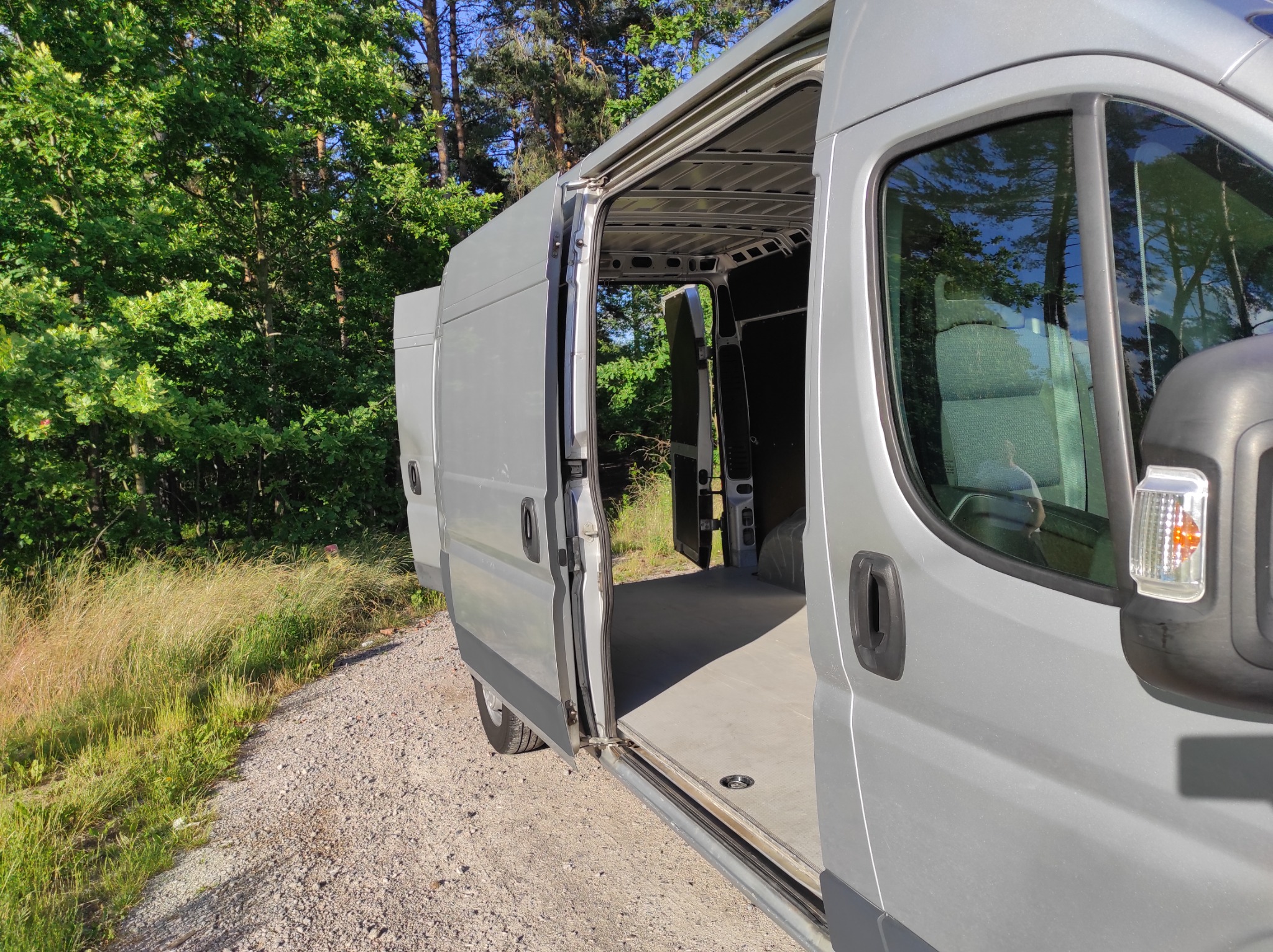 Srebrny bus dostawczy z otwartymi drzwiami bocznymi i tylnymi na żwirowej drodze, widok na puste wnętrze przestrzeni ładunkowej i otaczającą zieleń drzew.