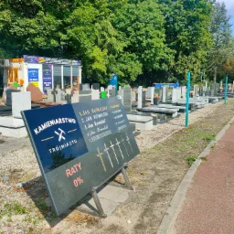 Ekspozycja nagrobków w zakładzie kamieniarskim, z tablicą reklamową oferującą raty 0% i przykładowymi nazwiskami z datami, na tle rzędu nagrobków i zieleni cmentarnej.