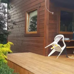 Częściowy widok brązowego domu z drewnianą elewacją, z przylegającym tarasem z drewnianych desek i schodkami; na tarasie odwrócone białe krzesło i stolik.
