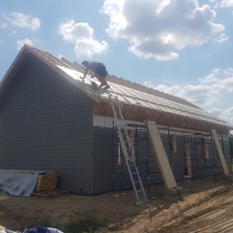 Twój Dom Szkieletowy - Budowa domu z szarym sidingiem: monter mocuje płyty na dachu, widoczne rusztowania, ułożone deski i niezagospodarowany teren wokół.