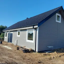 Szary dom jednorodzinny z ciemną dachówką, widok z zewnątrz na tle błękitnego nieba, teren wokół domu w trakcie prac ziemnych.