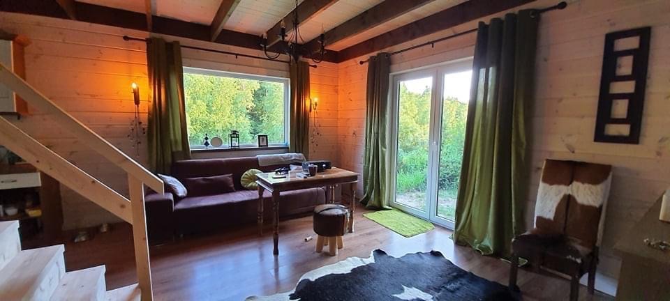 Wnętrze domu z drewnianych bali: salon z sofą, stolikiem, oknem z widokiem na zieleń, dekoracjami ściennymi i schodami.