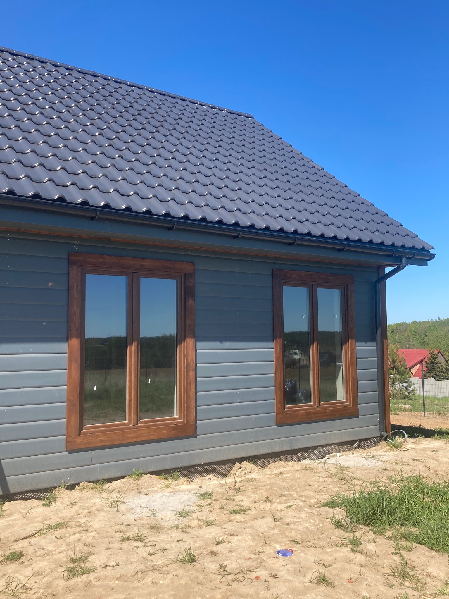 Niski, szary dom z dwoma oknami z drewnianymi ramami i ciemnoszarą dachówką, widoczny fragment elewacji i dachu, teren wokół domu w trakcie zagospodarowywania, z widoczną ziemią i fragmentami trawy...