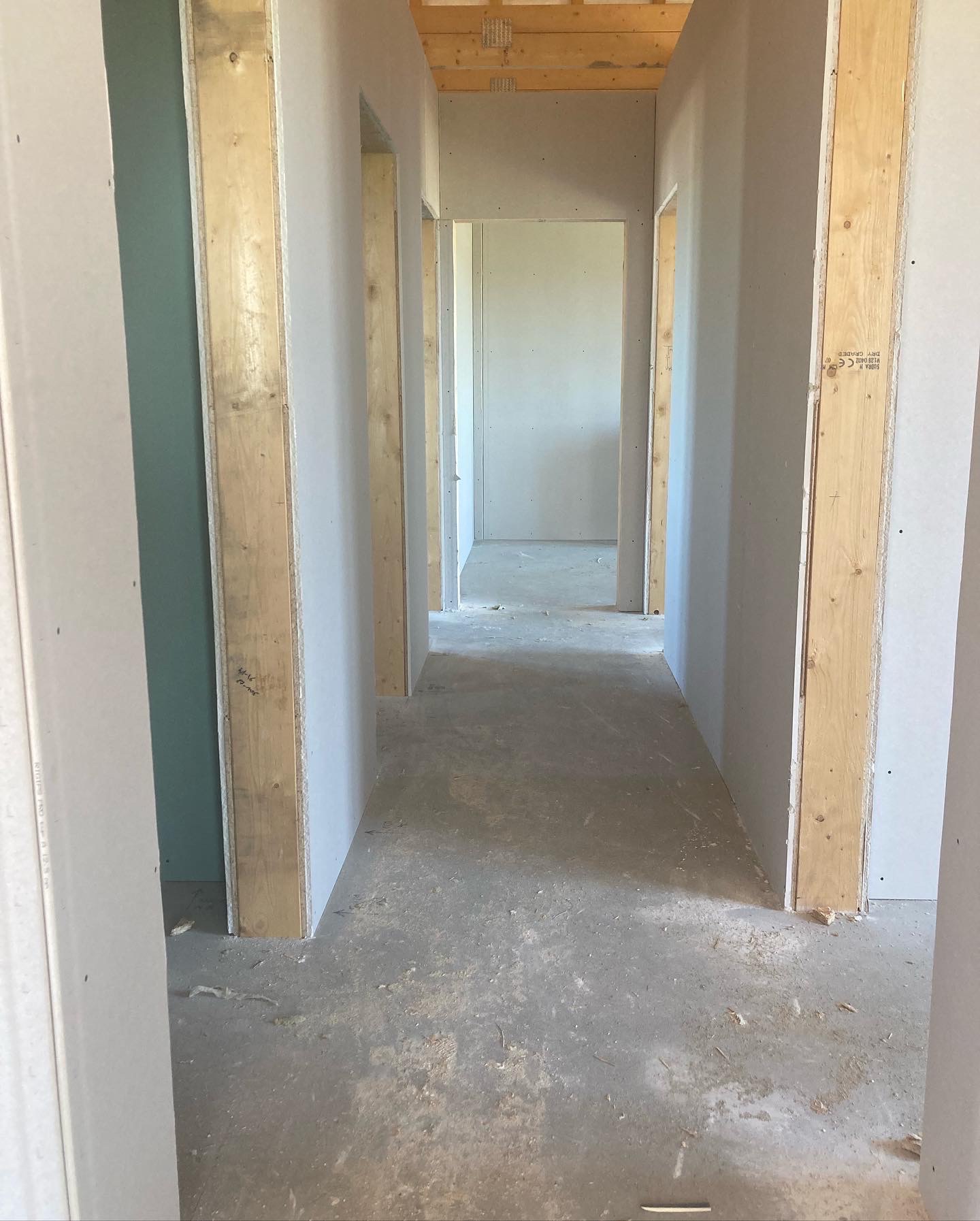 Wnętrze domu w trakcie budowy: surowe ściany z płyt gipsowo-kartonowych na drewnianej konstrukcji, widoczne drewniane belki stropowe, betonowa podłoga z resztkami materiałów budowlanych...