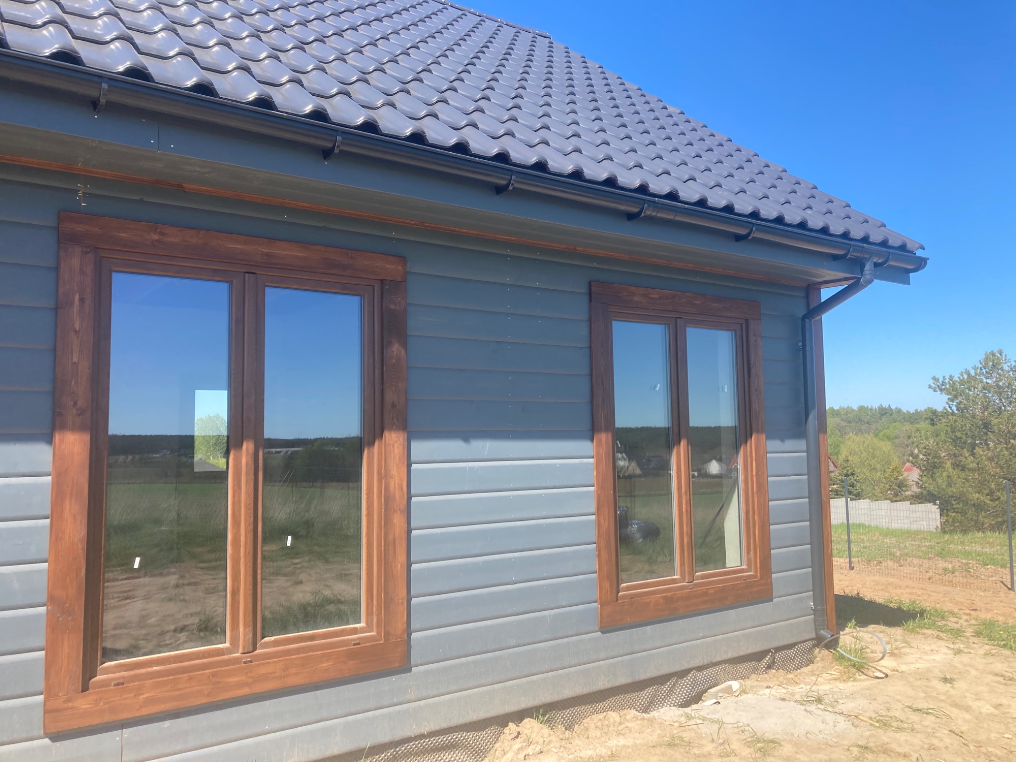 Fragment ściany zewnętrznej domu szkieletowego z dwoma oknami w drewnianych ramach, widoczne orynnowanie i fragment dachu z szarej dachówki, teren wokół domu w trakcie prac wykończeniowych.