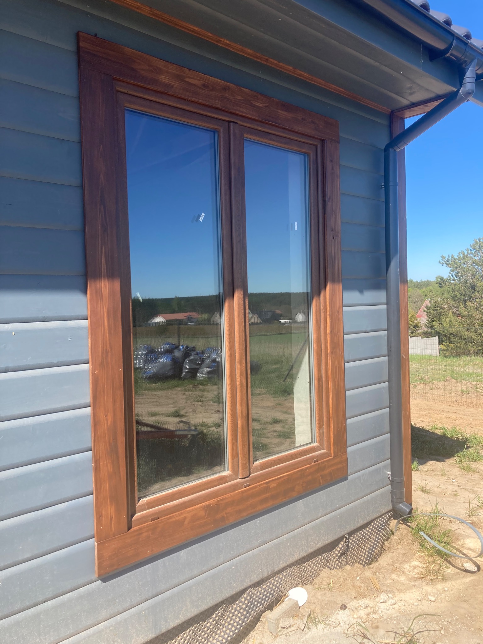 Fragment ściany zewnętrznej domu z drewnianą ramą okienną w kolorze orzecha i szarą elewacją z poziomych desek. Widoczny system rynnowy, grunt z geowłókniną i częściowo trawa.