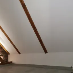 Poddasze po remoncie z drewnianymi belkami stropowymi, pomalowanymi ścianami na biało i szarą podłogą z płytek.