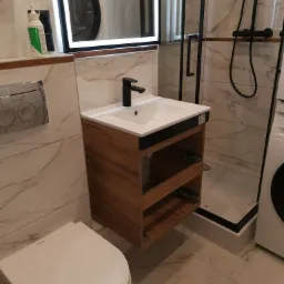 Nowoczesna łazienka z marmurowymi płytkami, wisząca szafka z drewnopodobnym wykończeniem, czarna armatura, kabina prysznicowa z czarnymi profilami i pralka widoczna w lustrze.