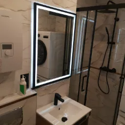 Nowoczesna łazienka z kabiną prysznicową z czarnymi profilami, umywalką z szafką z drewnopodobnym wykończeniem, lustrem z podświetleniem LED i pralką widoczną w odbiciu.