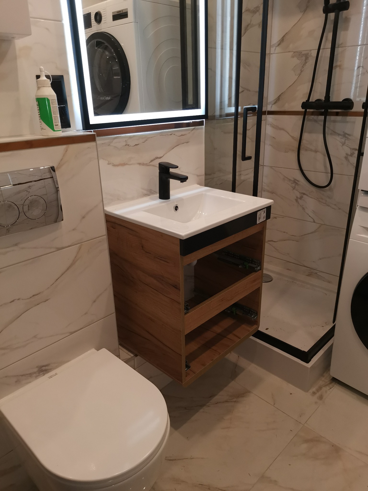 Nowoczesna łazienka z marmurowymi płytkami, wisząca szafka z drewnopodobnym wykończeniem, czarna armatura, kabina prysznicowa z czarnymi profilami i pralka widoczna w lustrze.