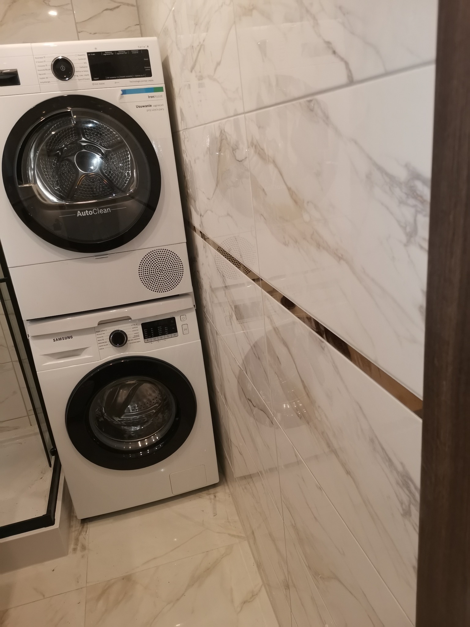 Ustawiona pionowo pralka i suszarka Samsung w łazience wykończonej marmurowymi płytkami z dekoracyjną, złotą listwą.