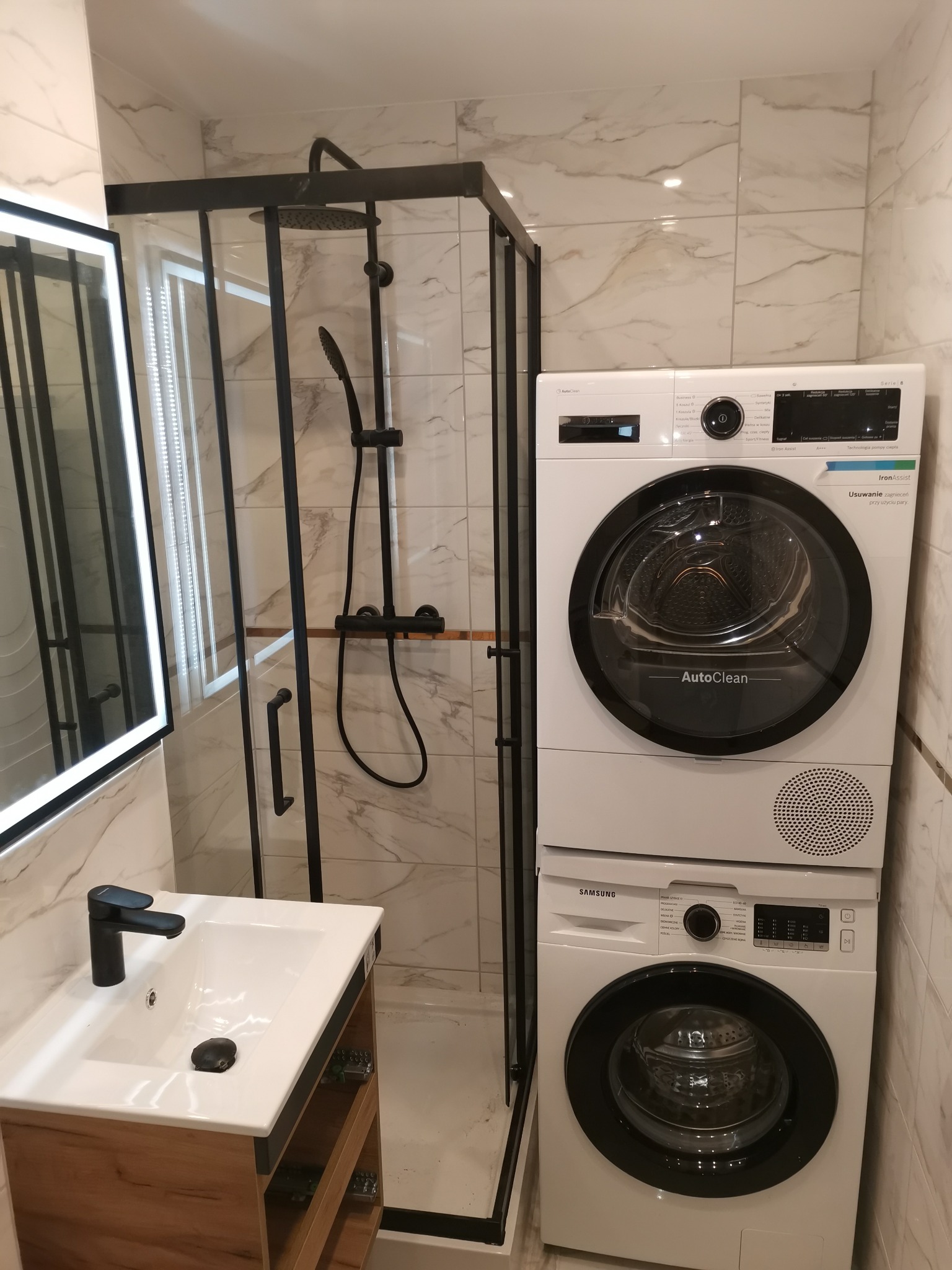 Nowoczesna łazienka z kabiną prysznicową typu walk-in z czarnymi ramami, umywalką nablatową z czarną baterią, marmurkowymi płytkami ściennymi i pralko-suszarką Samsung ustawioną w pionie.
